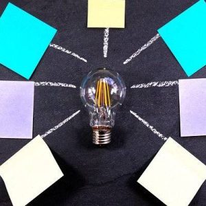 A lightbulb on a blackboard, with 7 chalk lines leading to 7 differently coloured blank post-it notes which are arranged in a circle around the lightbulb.