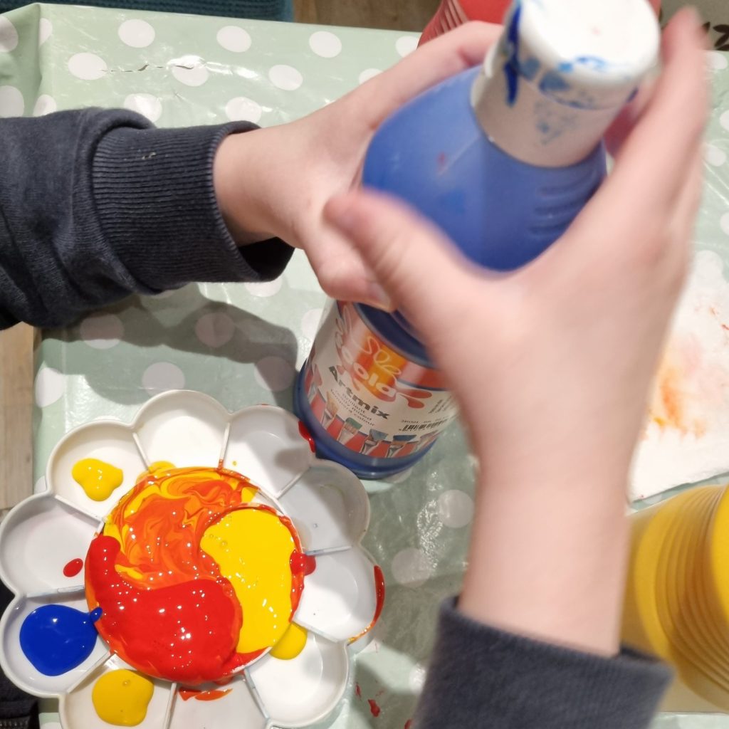 A child's hands hold a big bottle of blue paint. There is a paint palette filled with red, yellow and blue paint.
