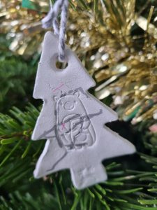 A home made clay ornament in the shape of a Christmas Tree, hanging on a Christmas Tree, with gold tinsel in the background. The ornament has a bear-shaped stamp and a silver scribble on.